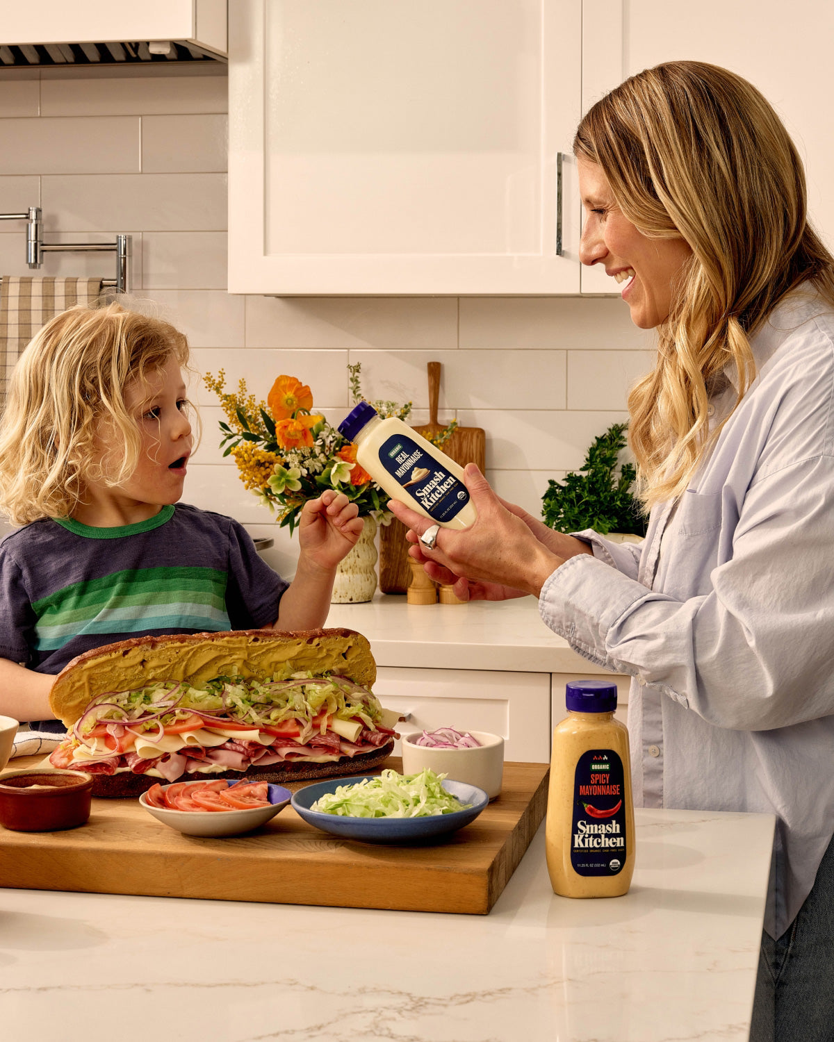A woman and child in the kitchen around a countertop with a large sandwich and Smash Kitchen products.