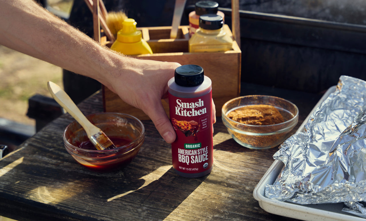 Hand reaching in to grab Smash Kitchen American Style BBQ Sauce from a tabletop near a smoker.