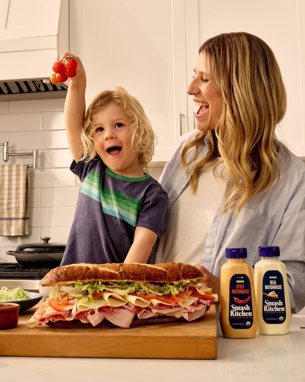 Woman and child in the kitchen with a large sandwich and Smash Kitchen products on the counter.